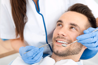Dentist wearing gloves examining a smiling man’s teeth with dental tools in clinic.