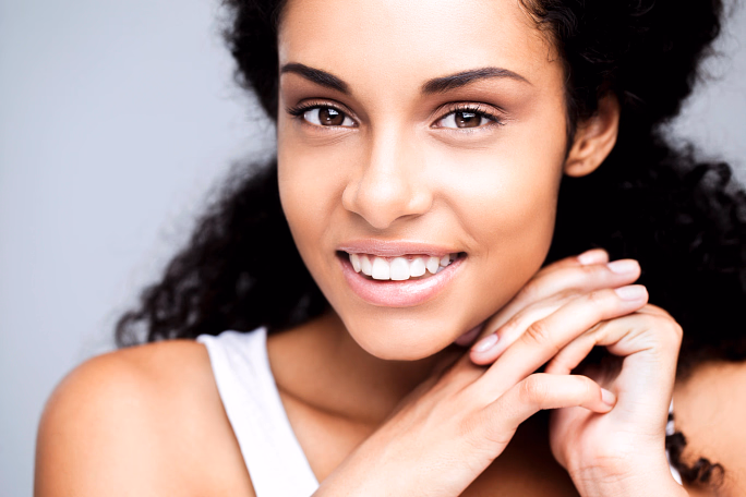 Close-up of a smiling woman with curly hair resting her chin on folded hands.
