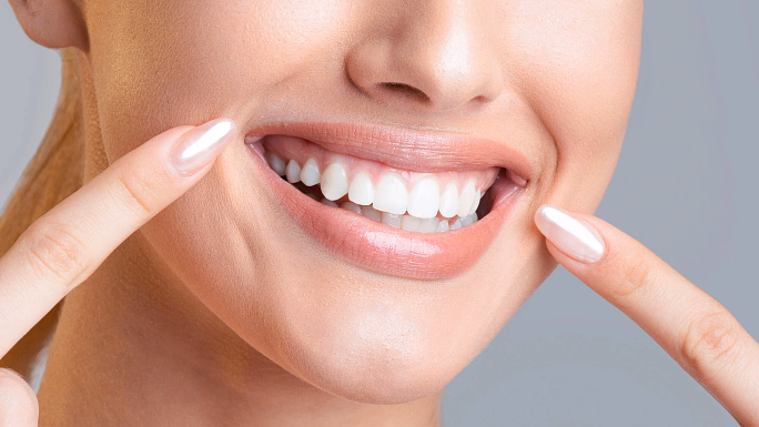 Close-up of a smiling woman pointing at her white teeth with manicured nails.
