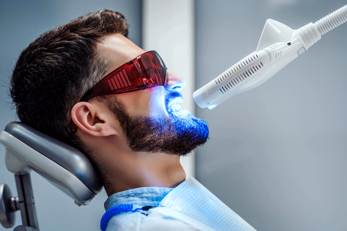 Man wearing protective glasses receiving blue light teeth whitening treatment in dental chair.