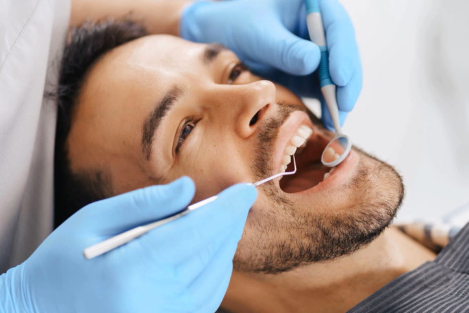Male patient smiling as dentist with blue gloves examines teeth with mirror and probe.