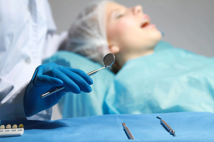 Dental professional wearing blue gloves holding a dental mirror near a patient covered in a blue drape.