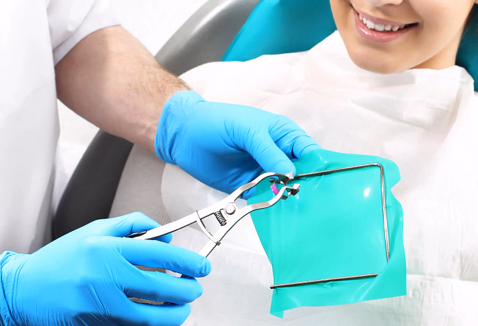 Dentist wearing blue gloves removing dental dam clamp near smiling patient's mouth.