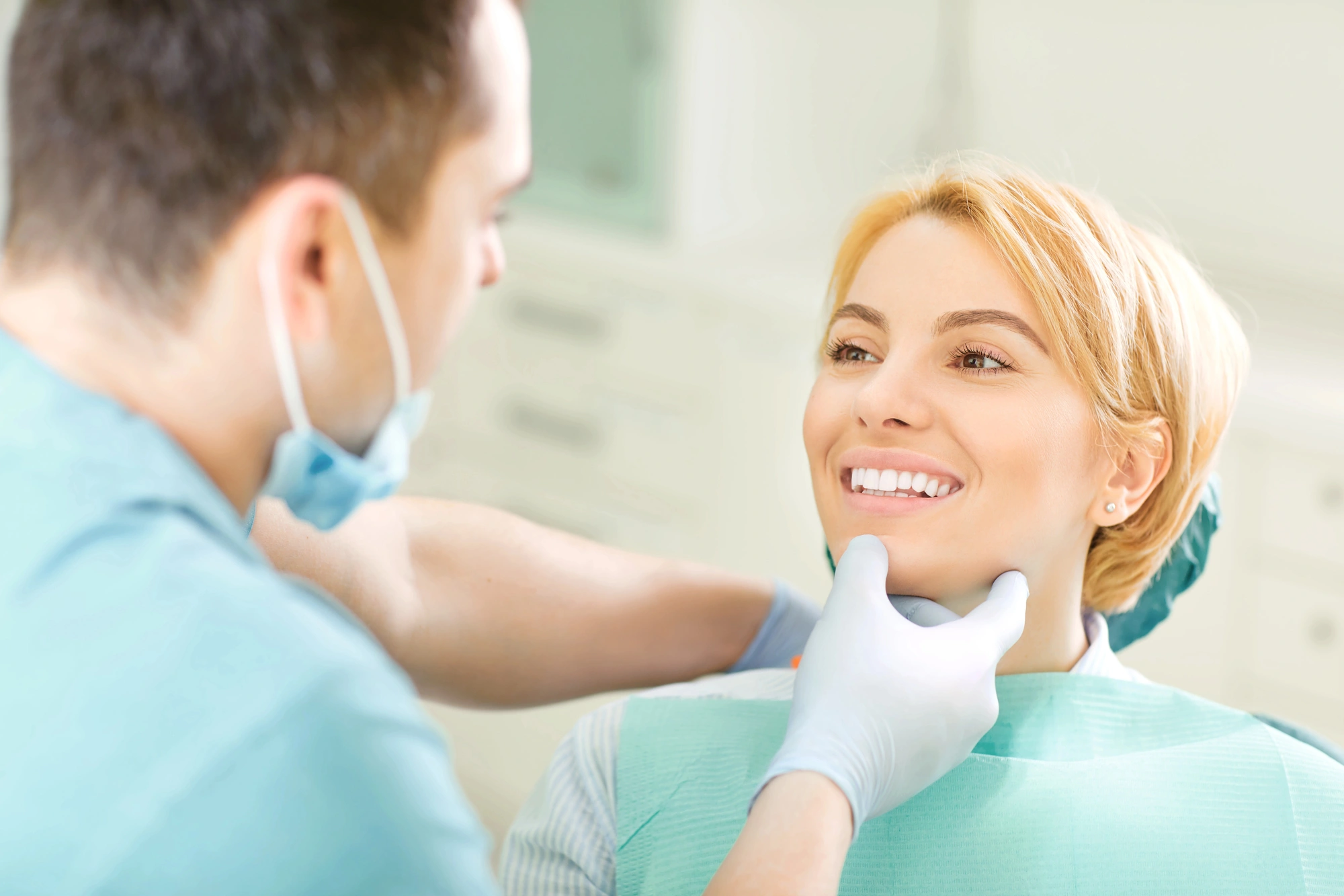 Dentist wearing gloves examines smiling female patient's teeth in dental office.