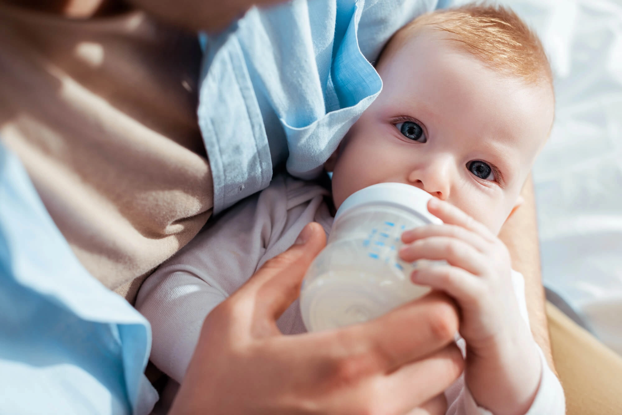 Close-up of a baby with blue eyes drinking milk from a baby bottle held by an adult.
