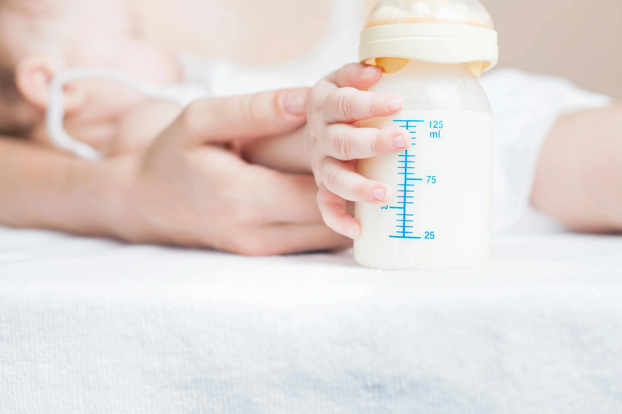 Baby's hand holding a milk-filled bottle on a white cloth with adult hand in the background.