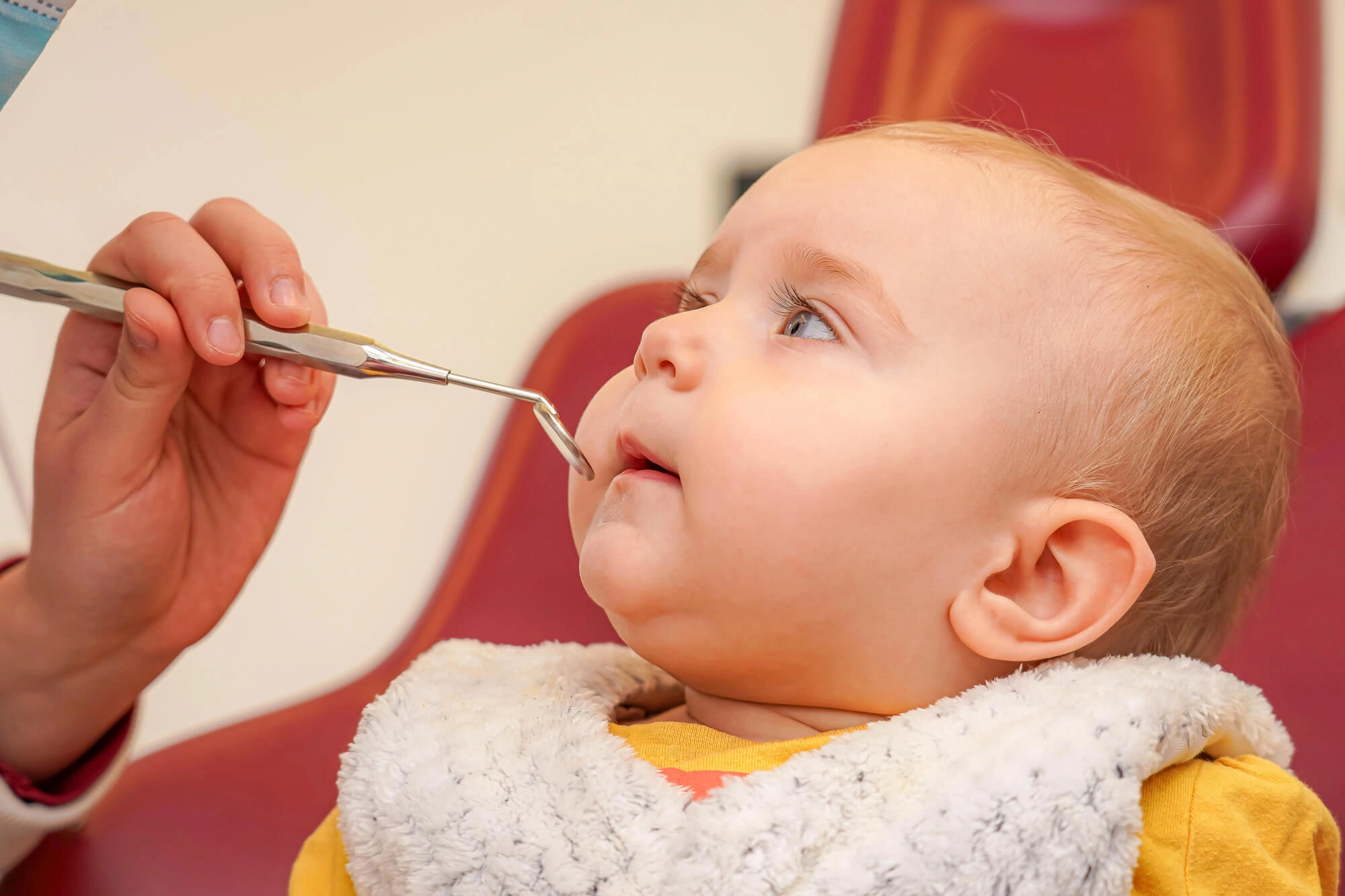Infant sitting on a red dental chair with mouth slightly open, a hand holding a dental mirror near mouth.