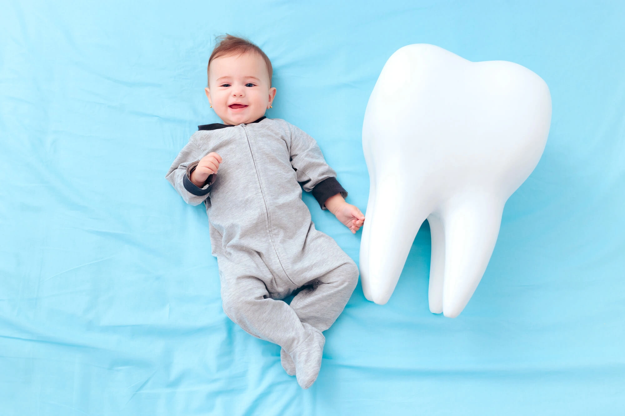 Smiling baby in grey onesie lying on blue sheet beside a large white model tooth.