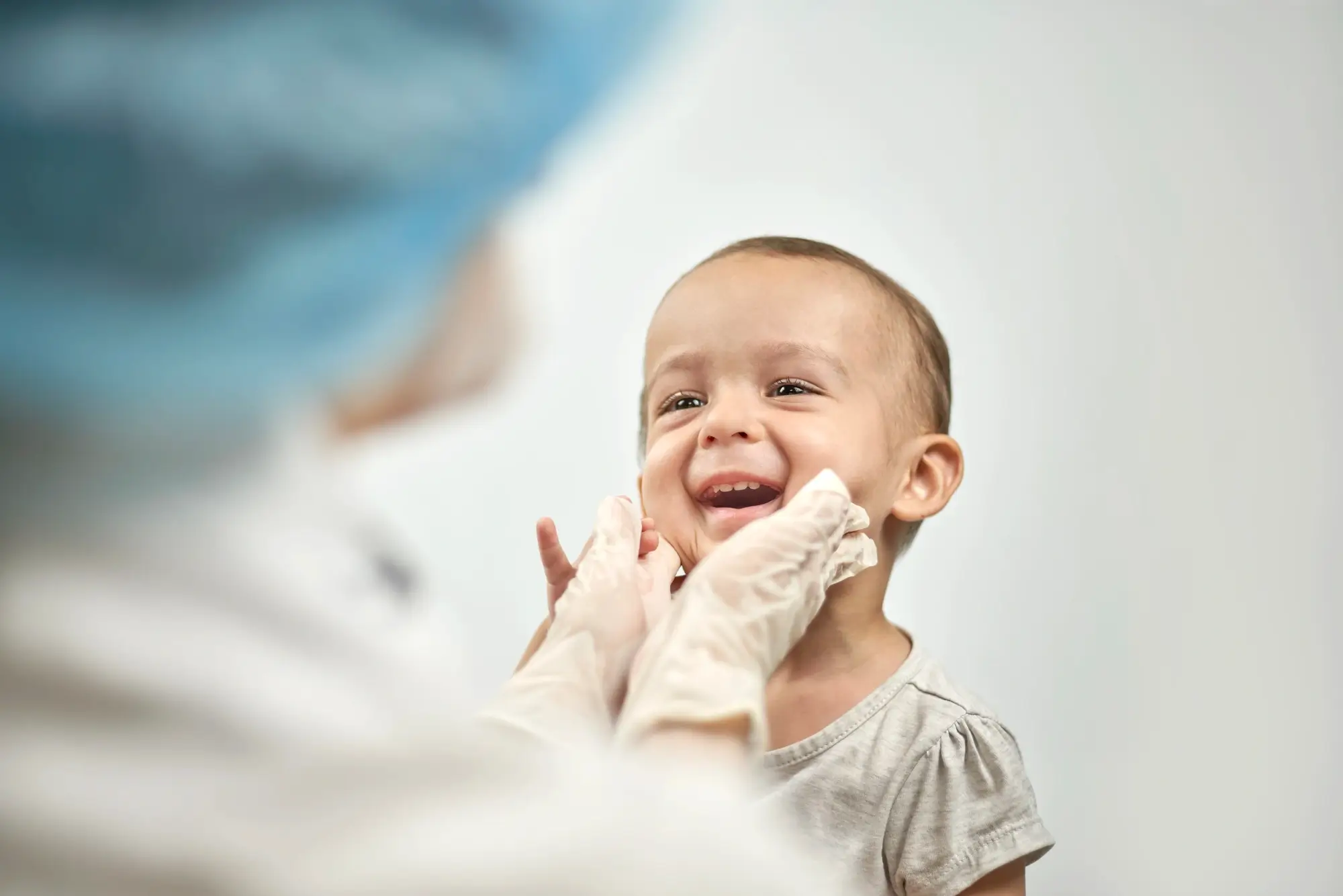 Smiling baby girl being gently examined by a healthcare professional wearing gloves and a blue cap.