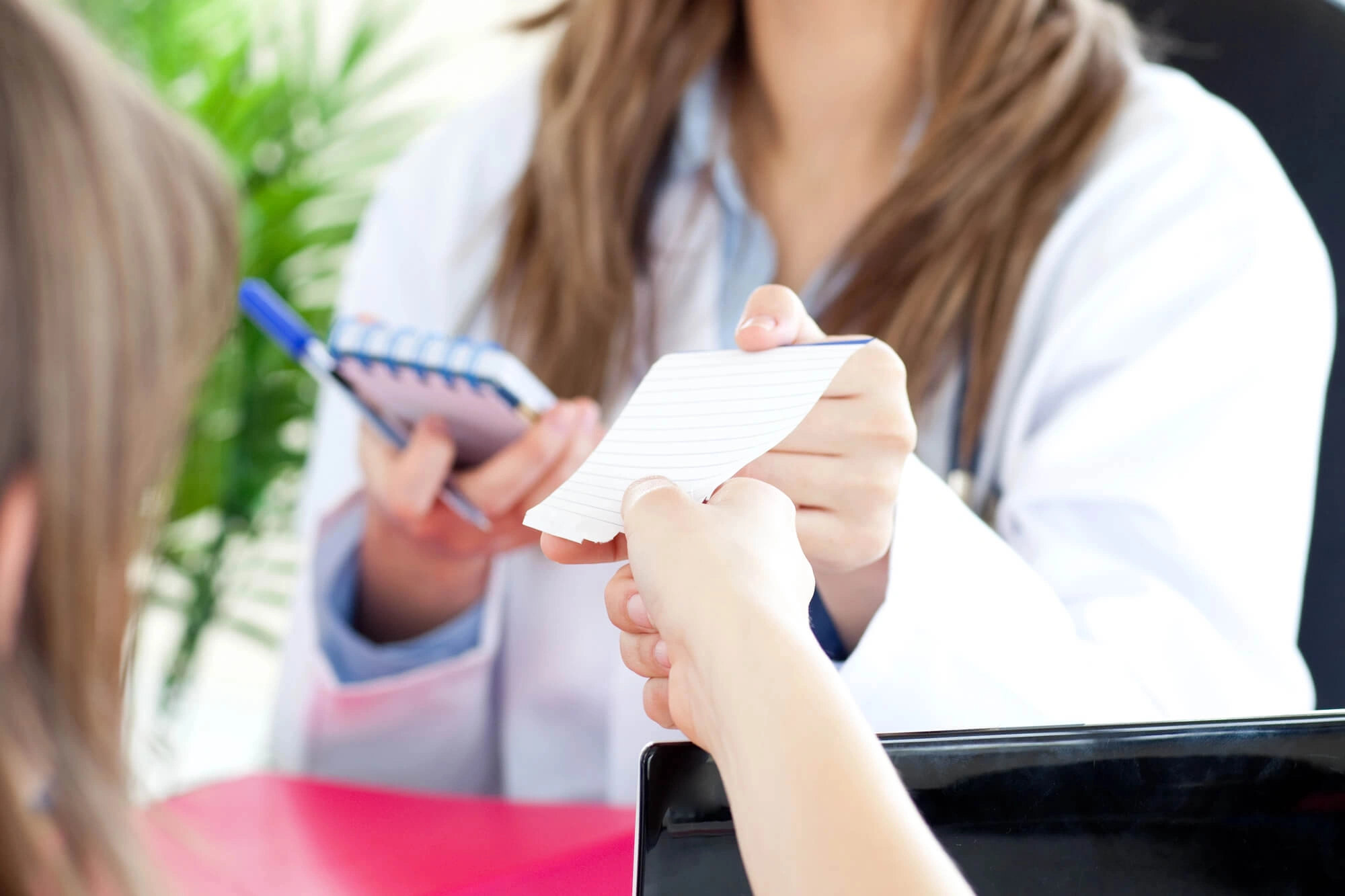 Doctor in white coat handing a prescription or note to a patient with a notebook and pen in hand.