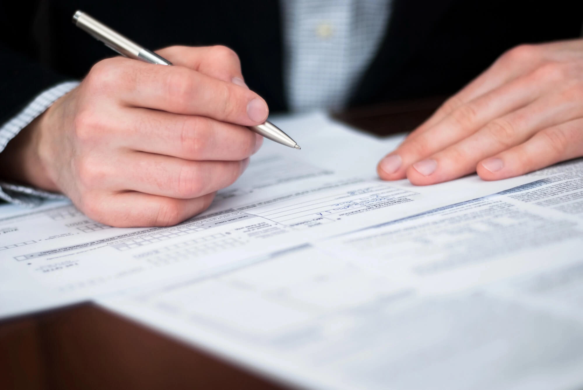 Person filling out printed forms with a silver pen on a desk.