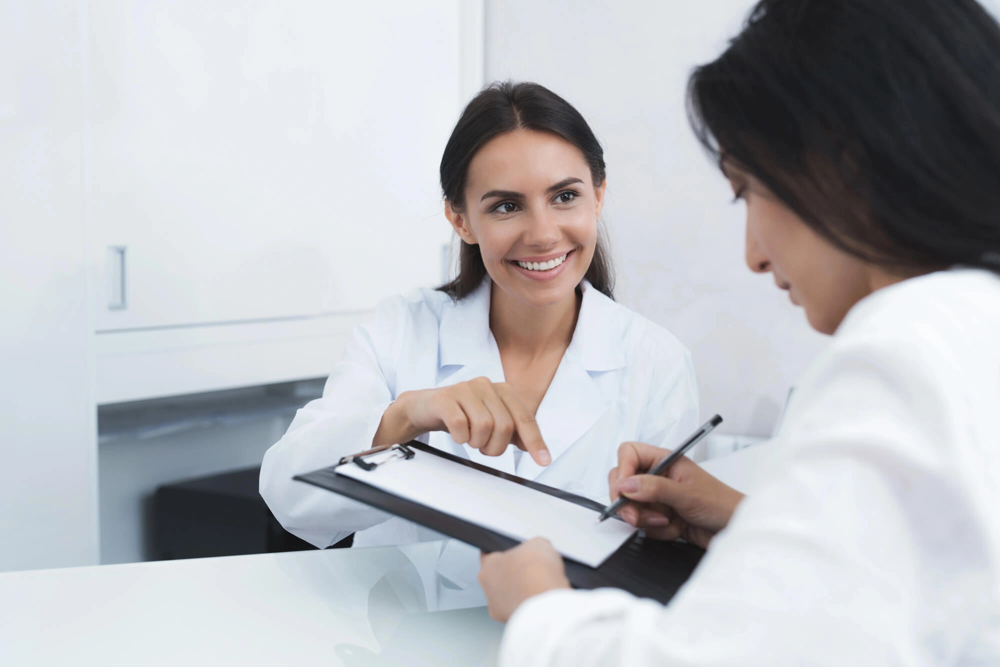 Smiling female doctor pointing to clipboard as patient signs a document in a medical office.