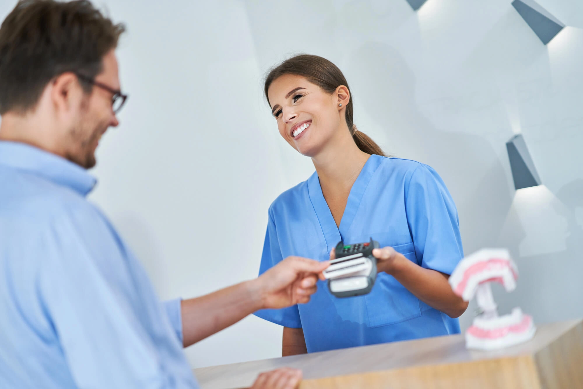 Smiling woman in blue scrubs holding card reader as man pays with credit card at reception.