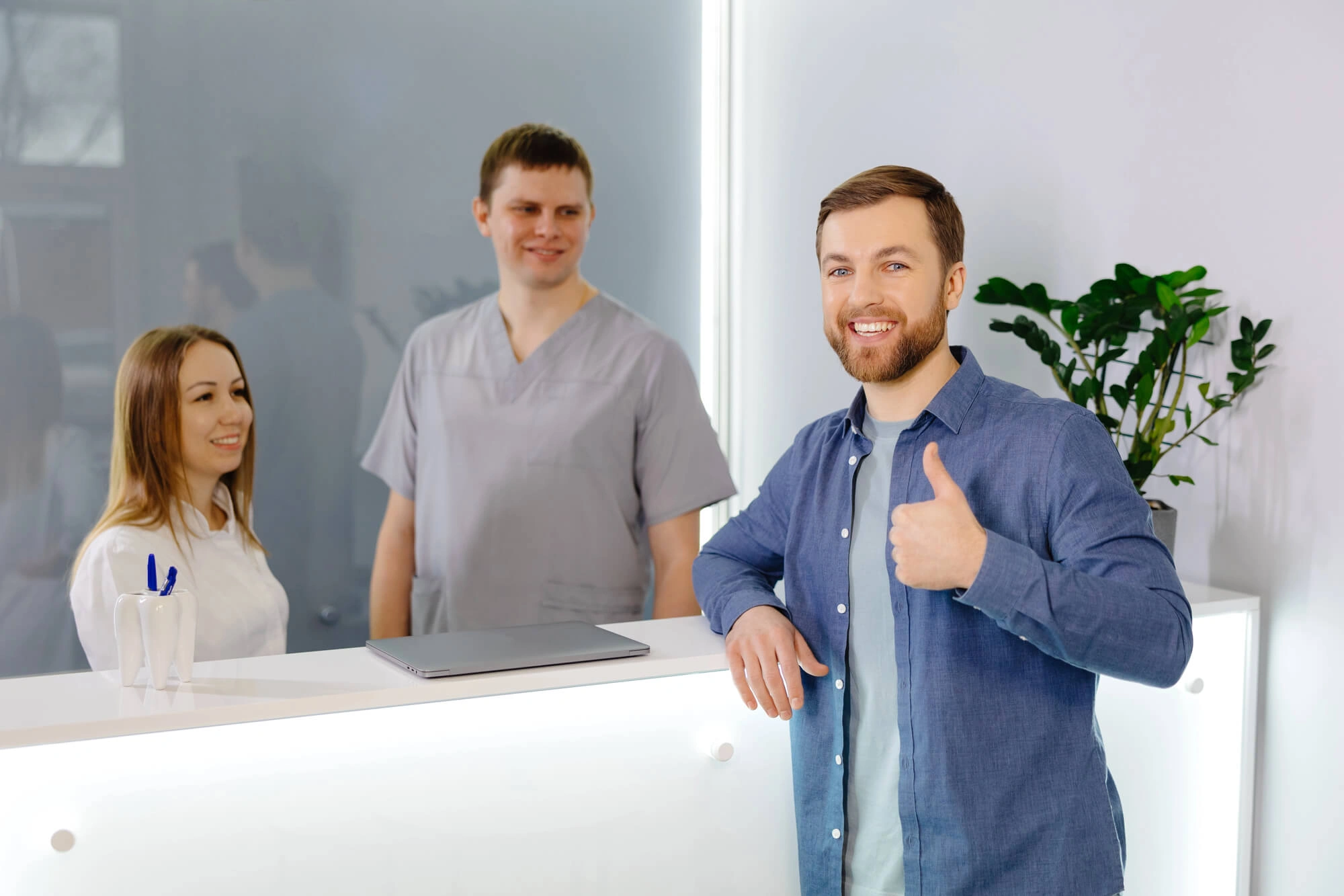 Smiling man giving thumbs up at a reception desk with two dental professionals behind it.