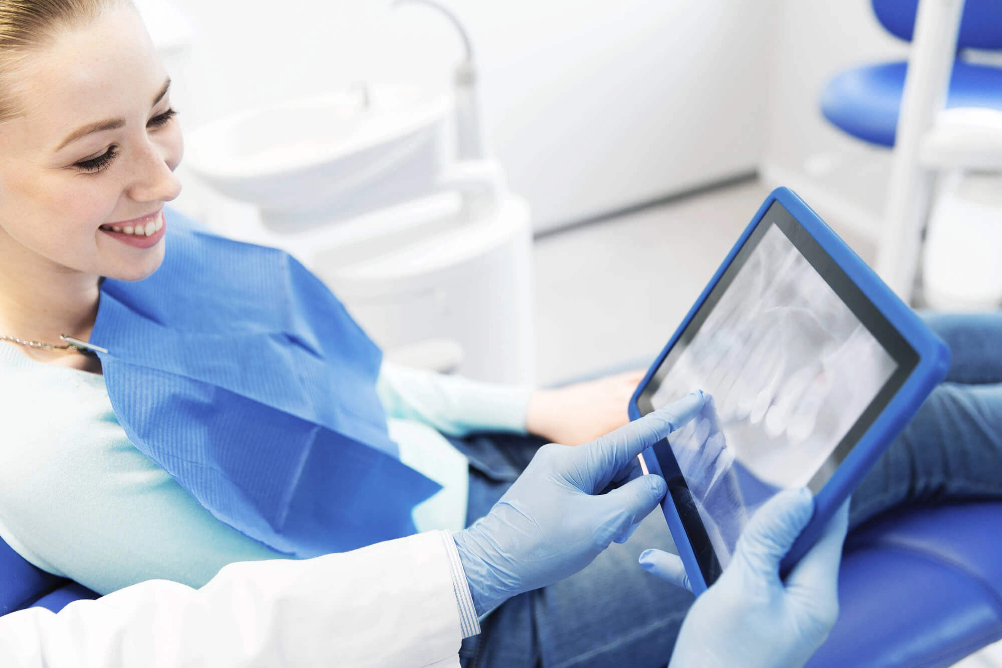 Dentist wearing gloves showing a dental X-ray on a tablet to a smiling female patient in a clinic.