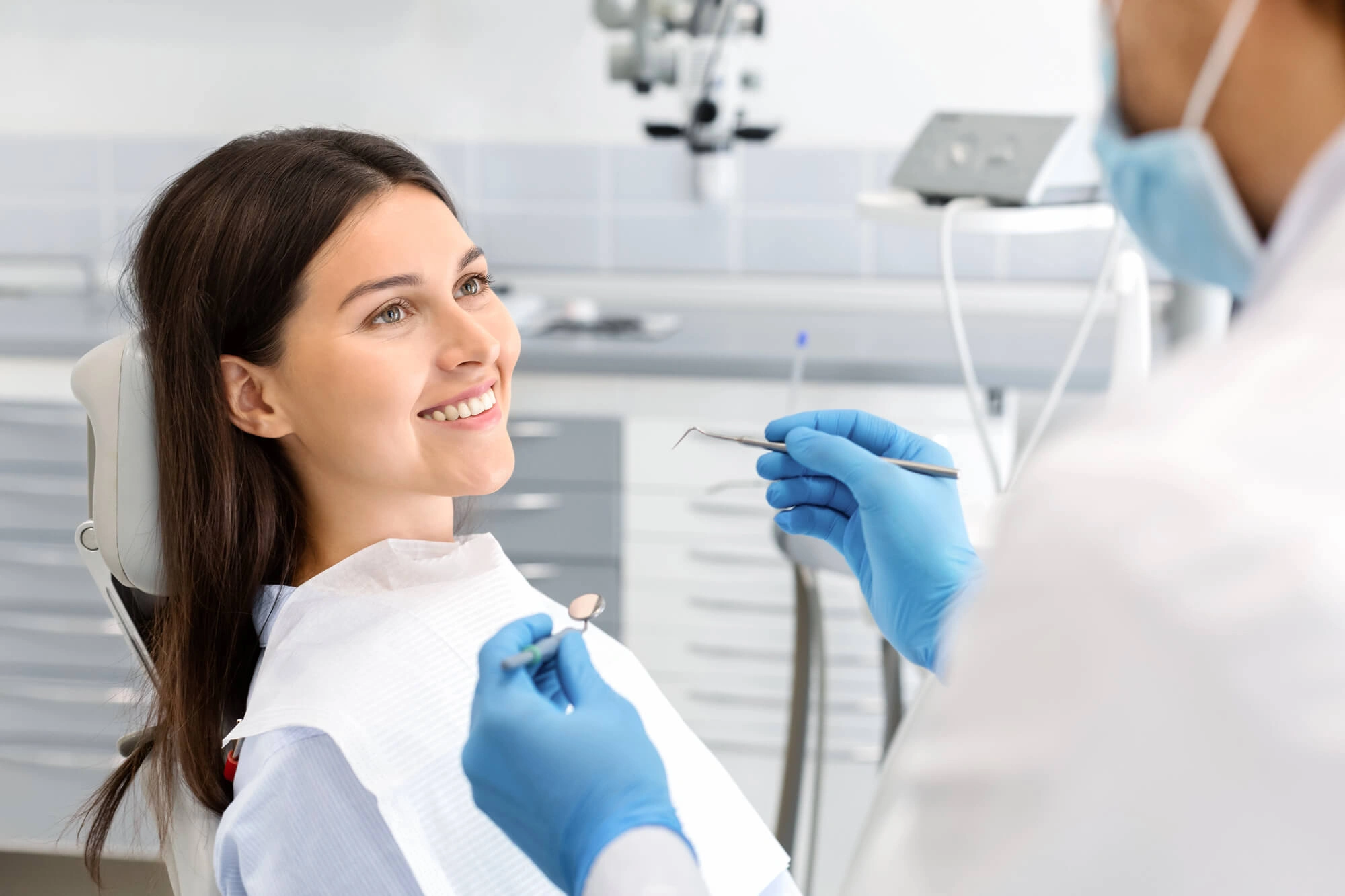 Smiling woman reclines in dental chair while dentist holds dental mirror and probe.
