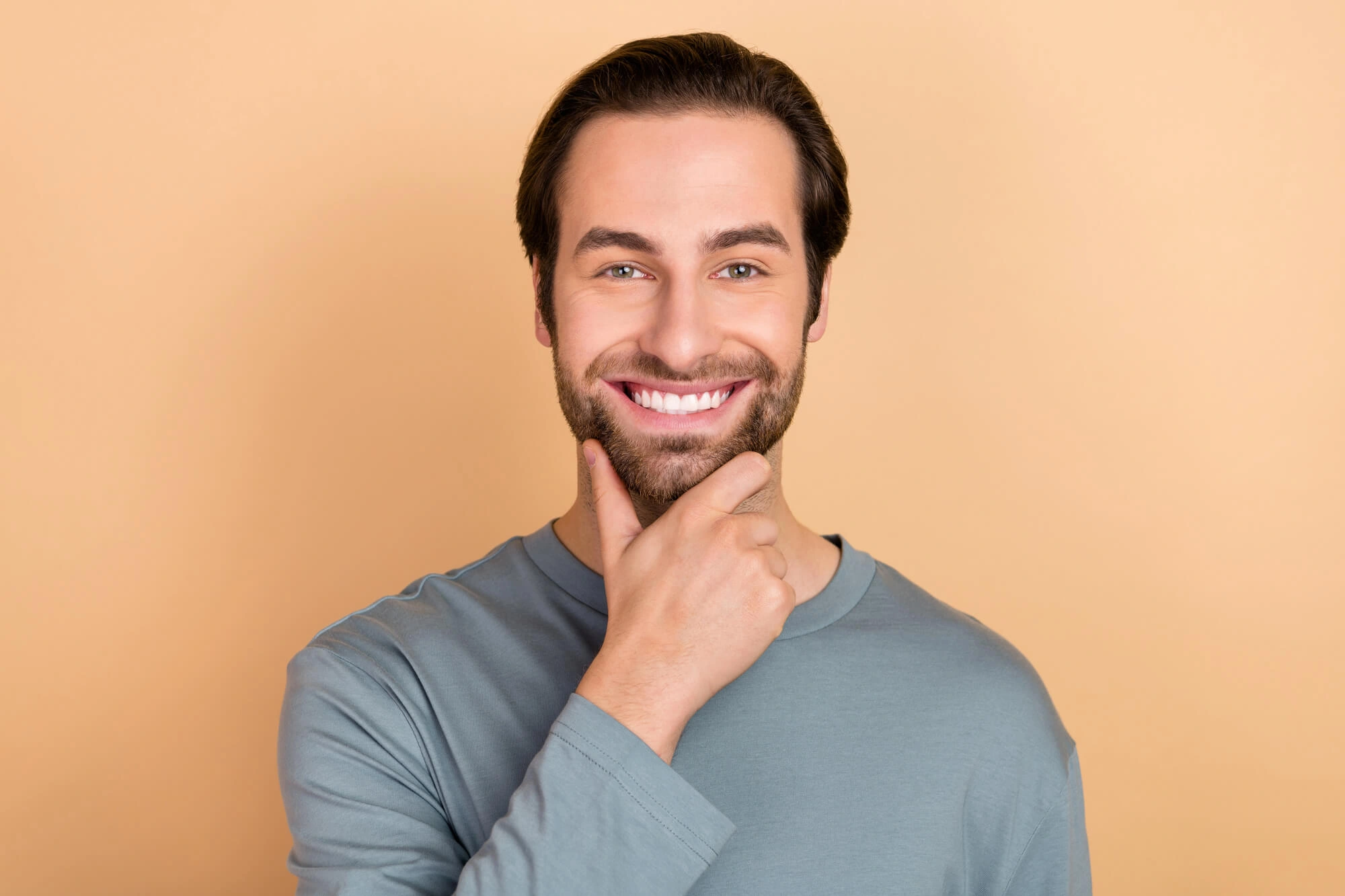 Smiling man with beard and blue shirt touching his chin against a beige background.