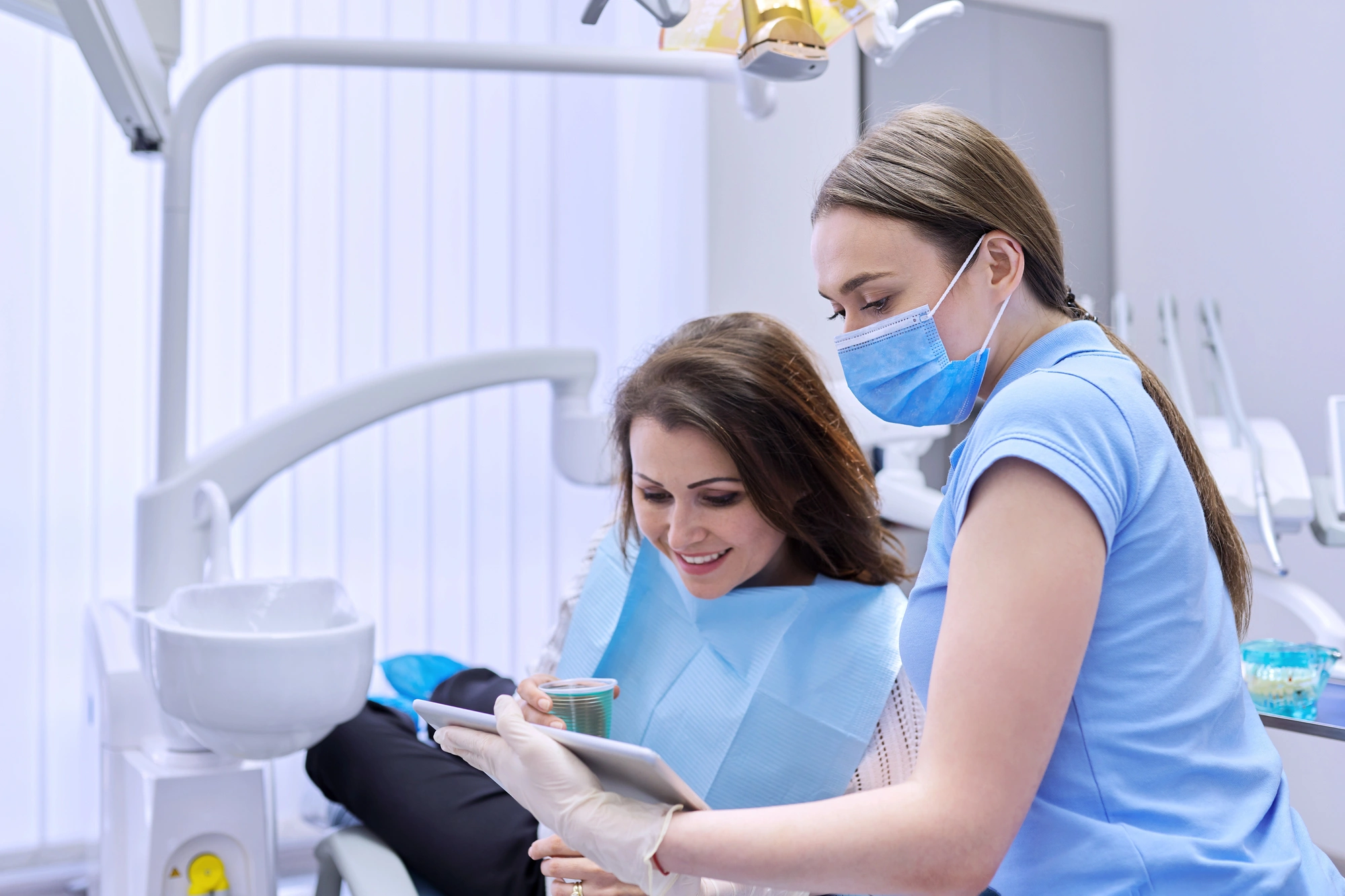 Dental assistant showing a tablet to a smiling patient seated in a dental chair wearing a blue bib.