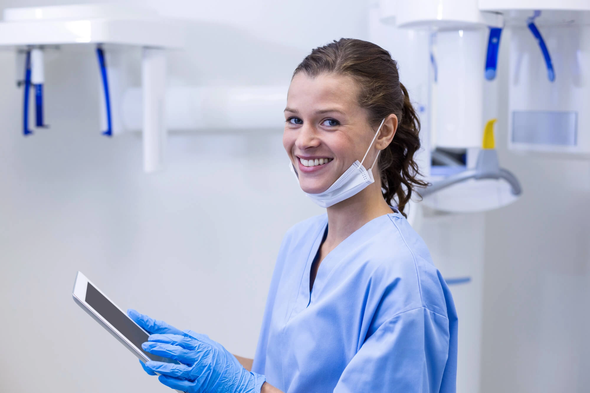 Smiling dental professional in blue scrubs and gloves holding a tablet in a dental clinic.