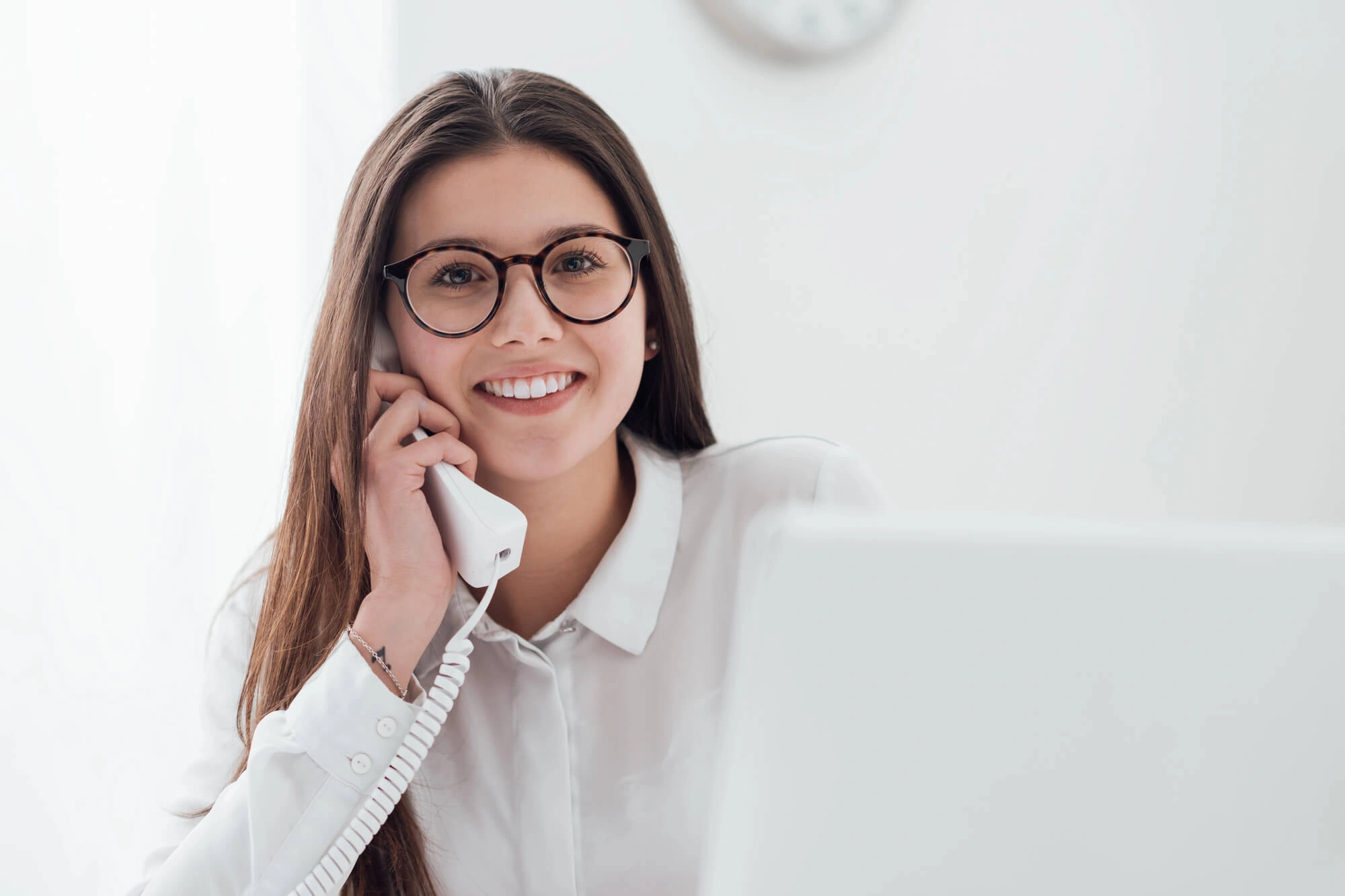 Smiling woman wearing glasses talking on a white corded phone in a bright office setting.