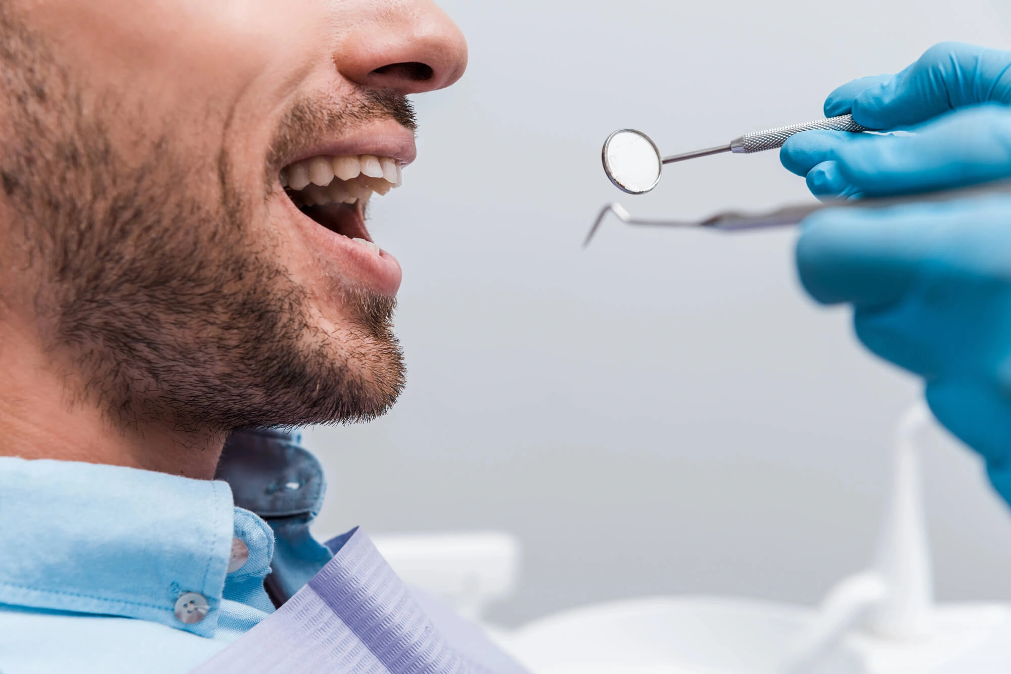 Man with beard opening mouth for dental exam with instruments held by gloved hands.
