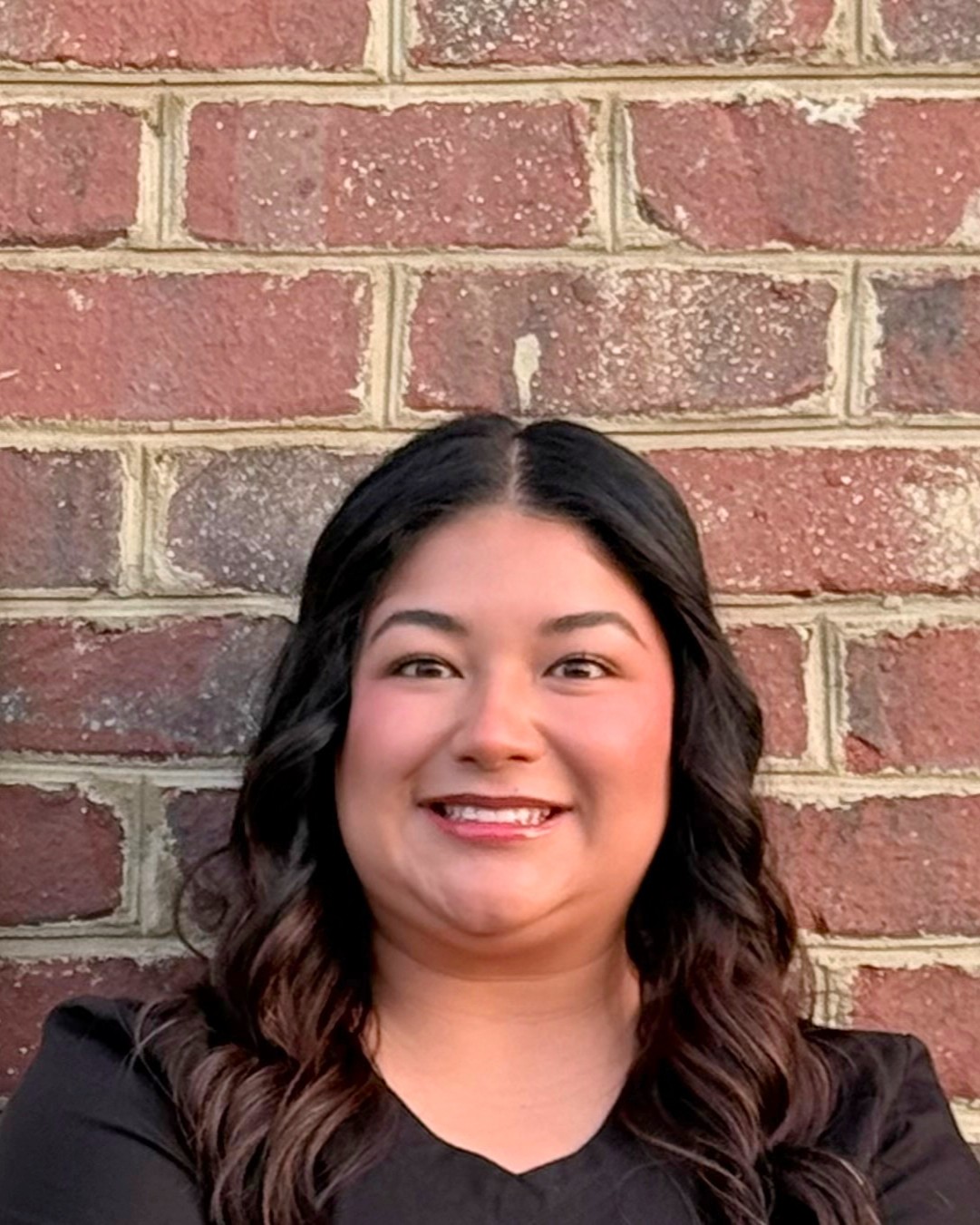 Smiling woman with dark wavy hair standing in front of a red brick wall.
