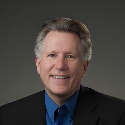 Smiling Doctor Grimes with gray hair wearing a blue shirt and black blazer against gray background.