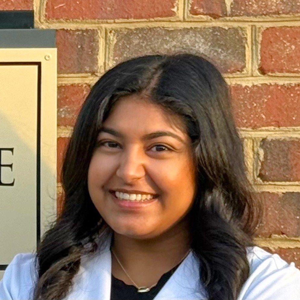 Smiling Doctor khal with long dark hair in white coat standing in front of a brick wall.