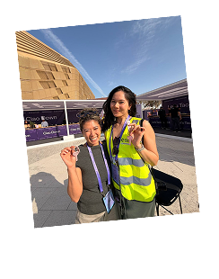 Two smiling women wearing lanyards and medals, one in a yellow safety vest, standing outdoors with buildings in the background under a clear sky.