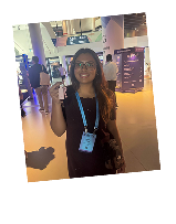 Smiling woman holding up a badge at a conference or event venue with people and displays in the background.