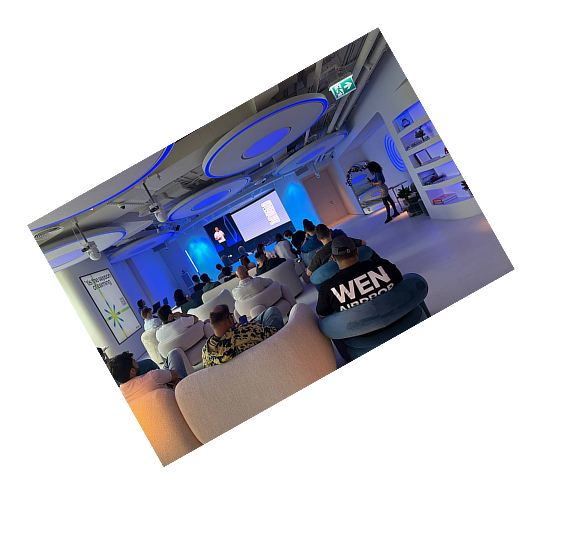 Audience seated on curved sofas and chairs watching a presenter and a screen in a modern conference room with blue lighting.