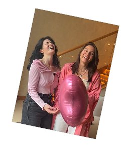 Two women laughing together indoors, one holding a large pink heart-shaped balloon.