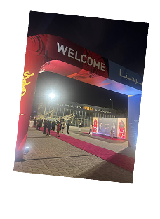 Nighttime event entrance with an illuminated red carpet and an archway displaying 'WELCOME' and Arabic text with people gathered in the background.