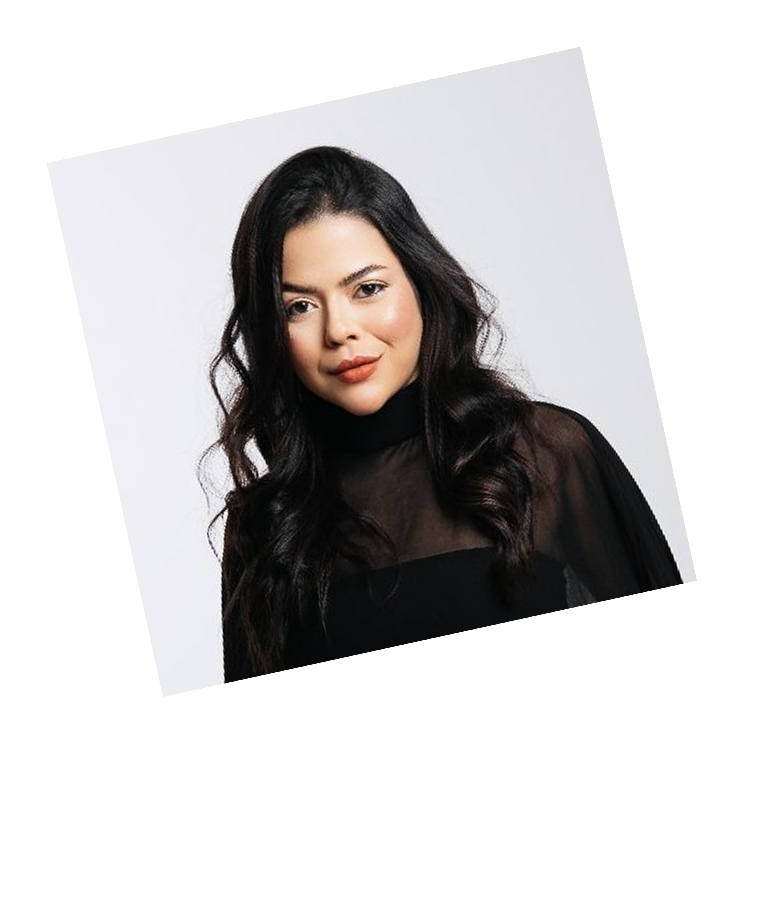 Portrait of a woman with long dark wavy hair wearing a black sheer top, smiling slightly against a light gray background.