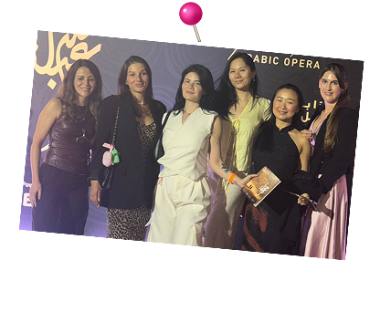 Six women standing together posing in front of a backdrop with Arabic Opera branding at an event.