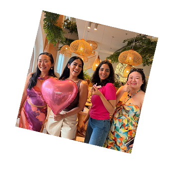Four smiling women standing indoors, one holding a pink heart-shaped balloon, with hanging lamps and green plants in the background.