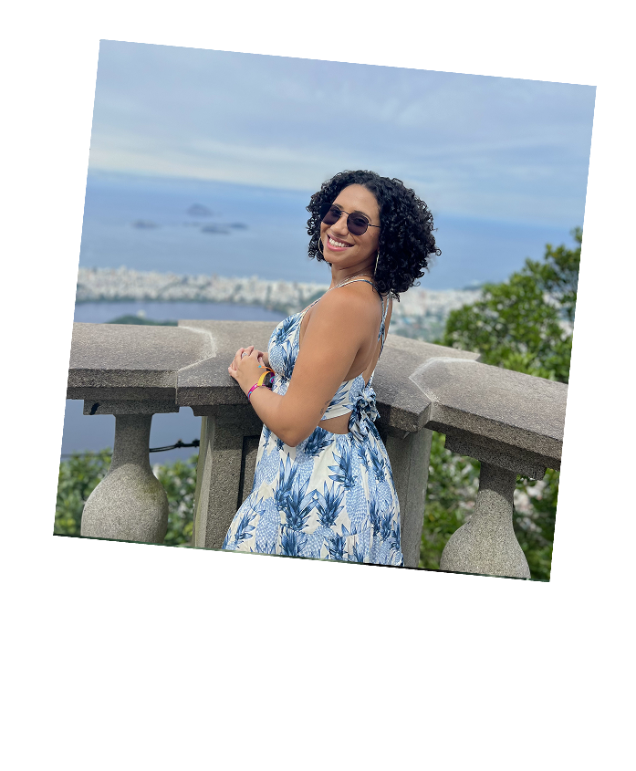 Smiling woman with curly hair wearing sunglasses and a blue and white dress stands by a stone railing overlooking a city and ocean view.
