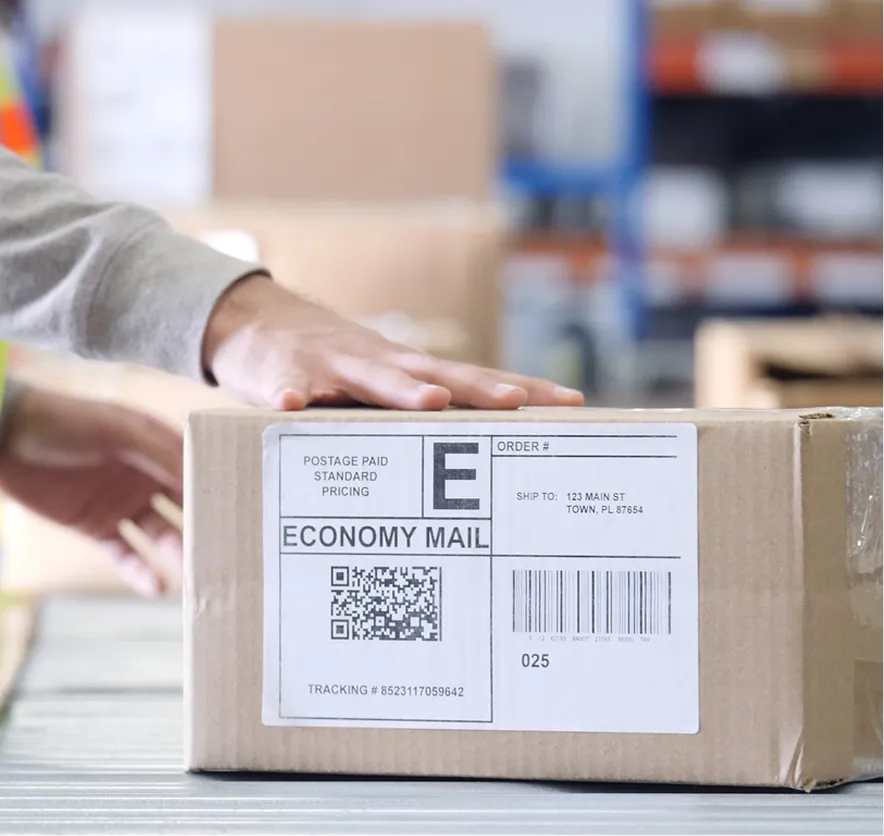 Hand placing a cardboard box with an economy mail shipping label on a conveyor belt in a warehouse.