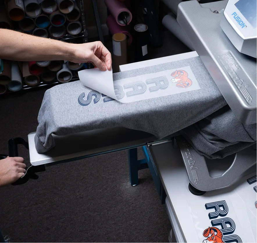 Hand peeling off a heat transfer print with the word 'RAD' and a tiger graphic from a gray t-shirt on a heat press machine.