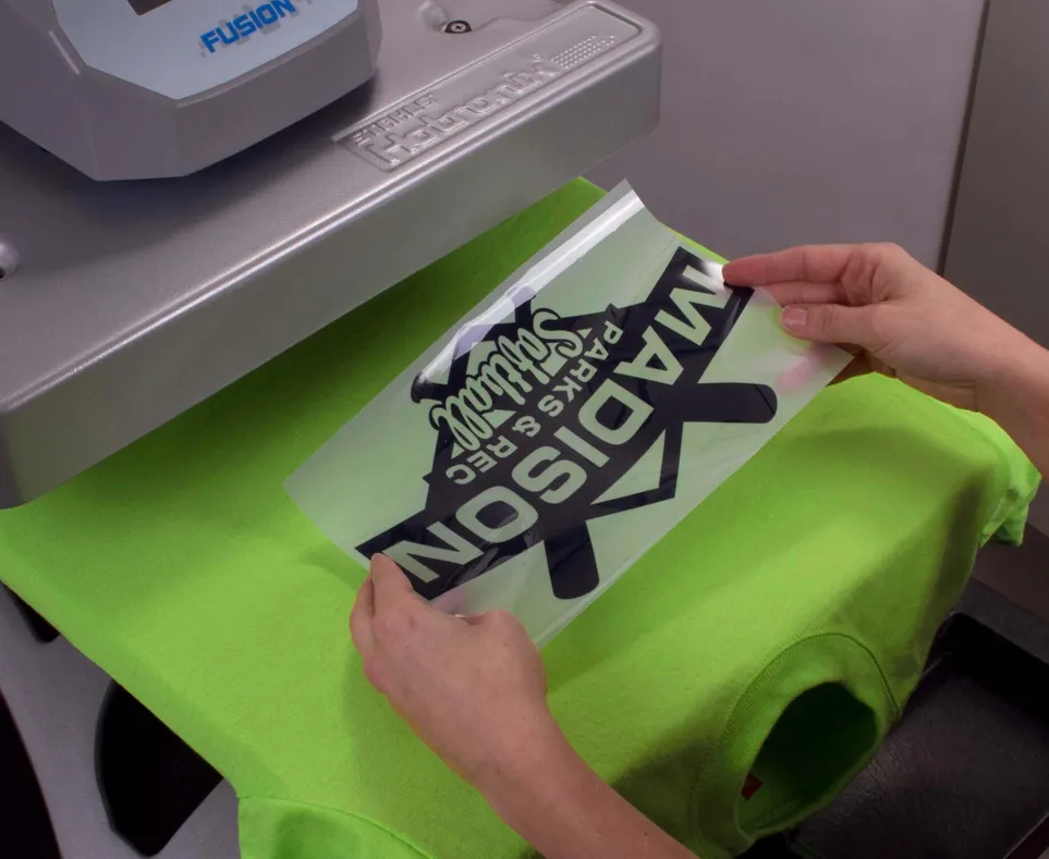 Hands holding a heat transfer vinyl sheet with 'MADISON Parks & Rec Salvage' text above a bright green shirt on a heat press machine.