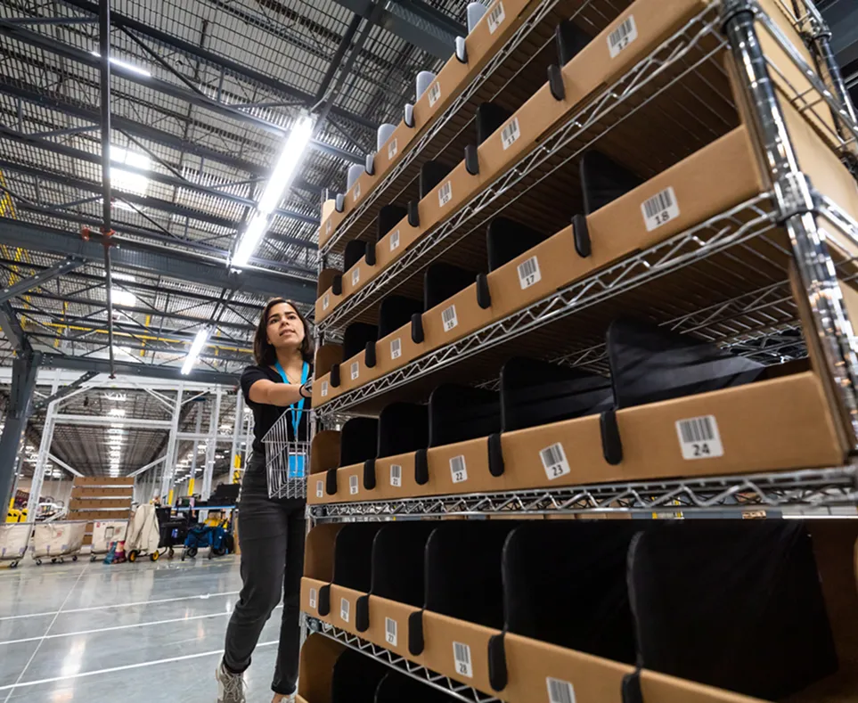 Worker organizing labeled bins on metal shelves inside a large warehouse with high ceilings and bright lighting.