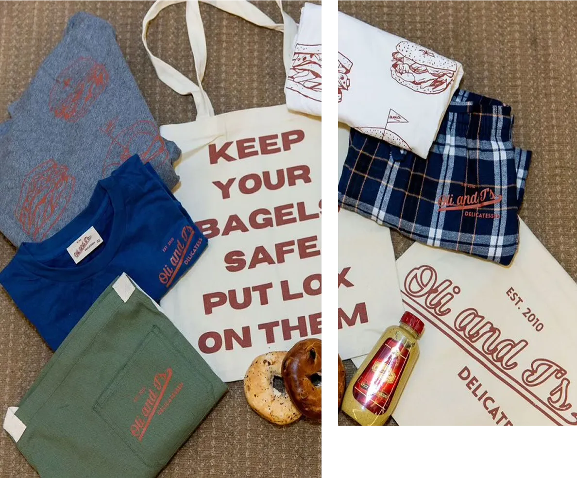 Assorted merchandise including t-shirts, tote bags with 'KEEP YOUR BAGELS SAFE PUT LOCK ON THEM' text, plaid shorts, two bagels, and a jar of mustard laid on a brown surface.