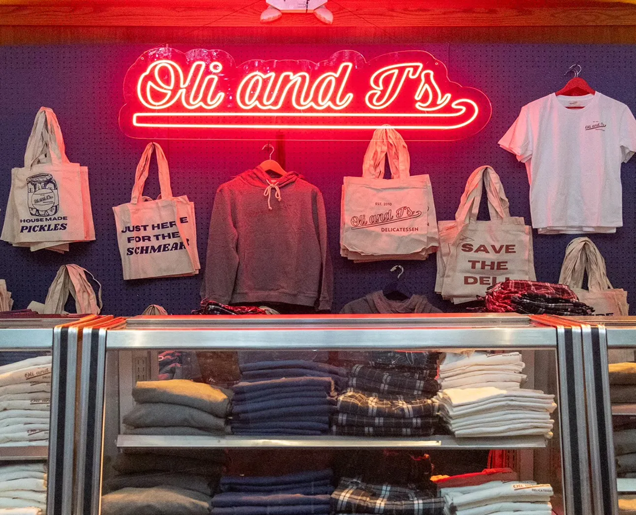 Retail display with a neon sign reading 'Oli and Ts', featuring tote bags, a hoodie, a t-shirt, and folded clothes beneath the display.