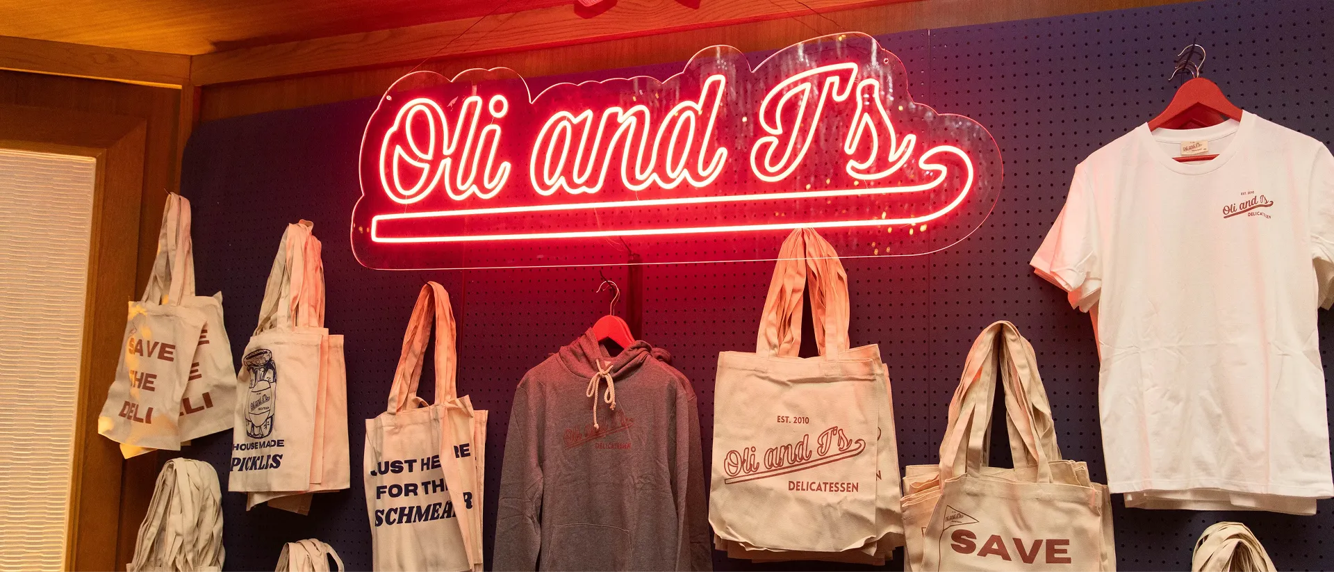 Display of branded merchandise including tote bags, a hoodie, and a white t-shirt under a red neon sign reading 'Oli and Ts'.