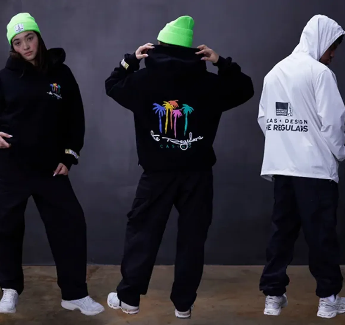 Three people wearing streetwear with black pants and white sneakers; two in black hoodies with colorful palm tree designs and one in a white jacket with text on back.