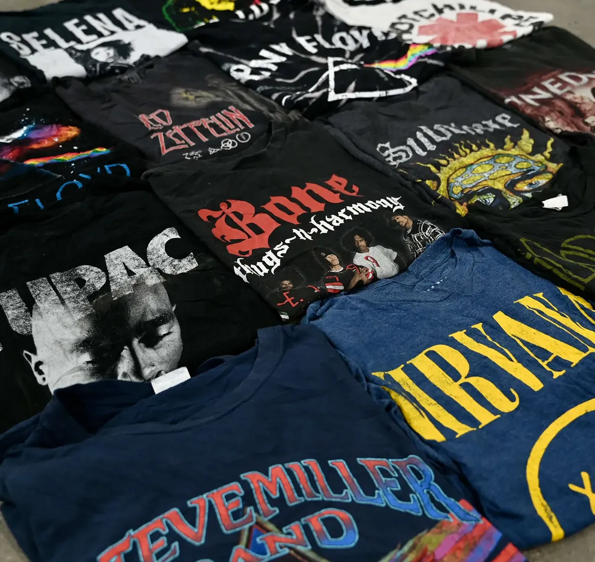 Collection of black and blue band t-shirts featuring artists like Nirvana, Tupac, Pink Floyd, Led Zeppelin, Bone Thugs-n-Harmony, and Steve Miller Band laid out on a surface.