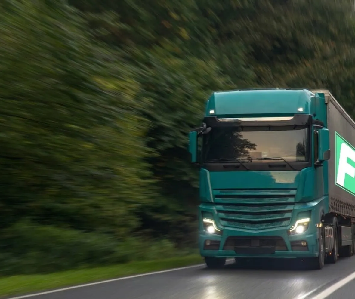 Green semi-truck with headlights on driving on a wet road surrounded by dense green trees.