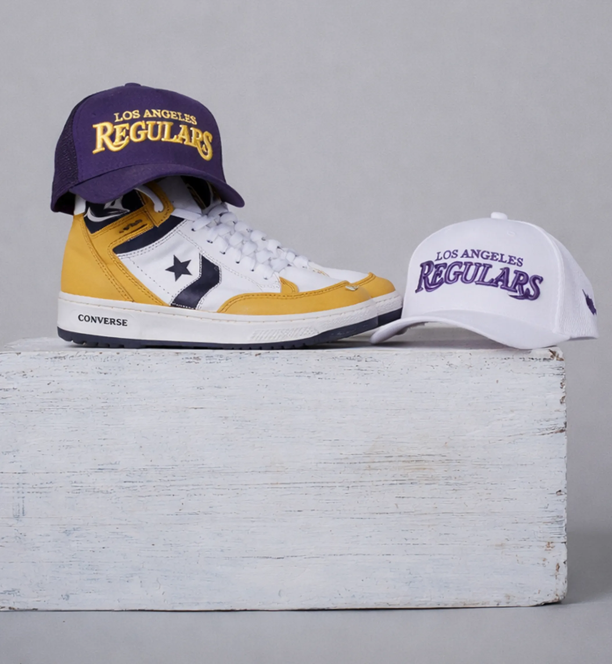 Pair of white and yellow Converse sneakers with a purple Los Angeles Regulars cap on top and a white Los Angeles Regulars cap beside them on a white wooden block.