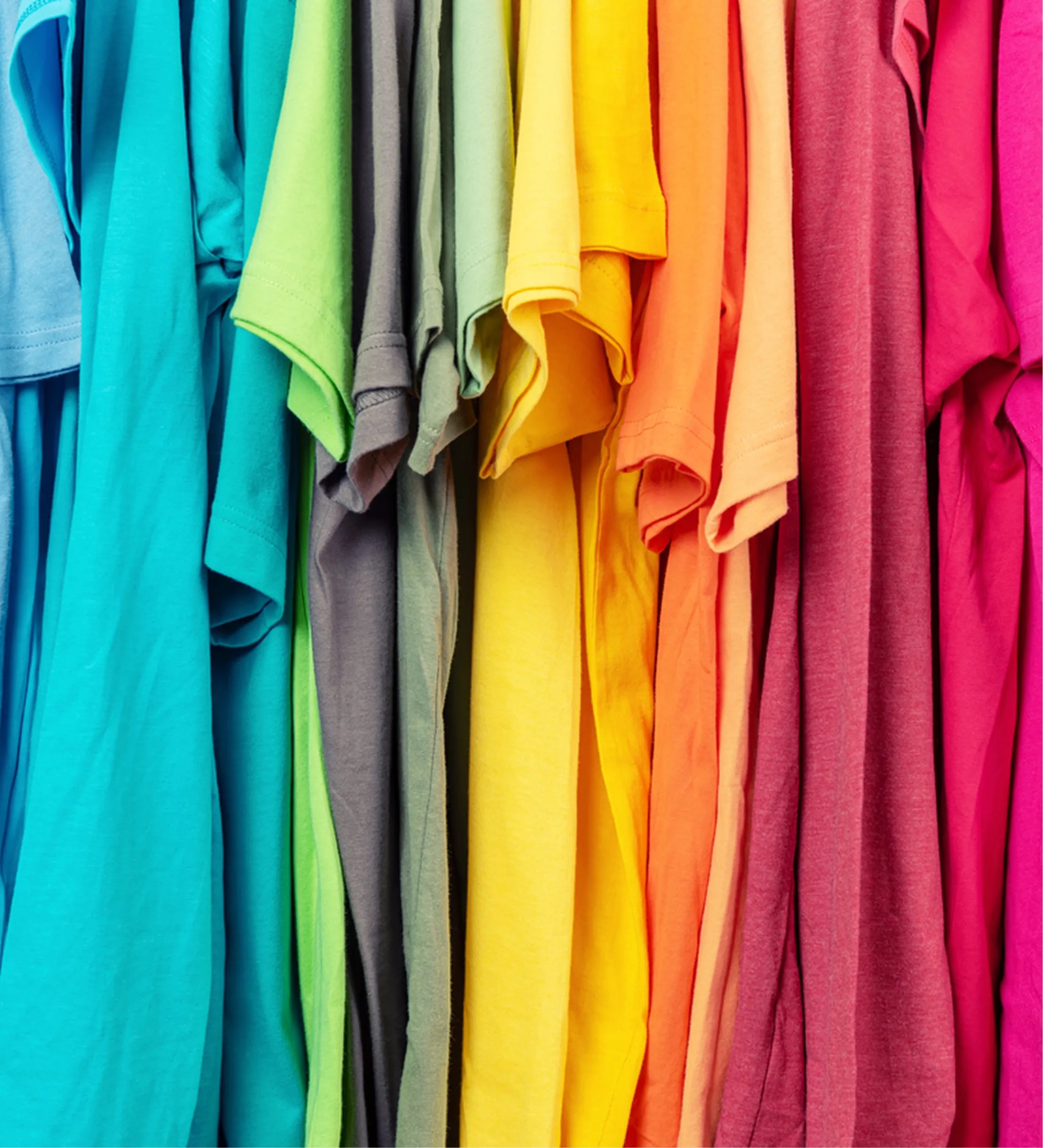 Rows of colorful t-shirts hanging closely with colors transitioning from blue and green to yellow, orange, and pink.