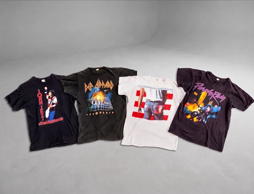 Four vintage rock band t-shirts laid out on a gray surface, featuring Jon Bon Jovi, Def Leppard, and Prince.